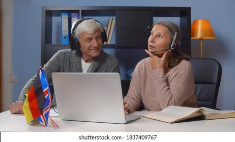 Aged Spanish And English Teachers In Headphones Giving Online Lesson. Portrait Of Senior Couple In Earphones Learning Foreign Language Online Watching Webinar On Laptop