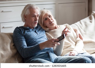 Aged Surprised Married Couple Embracing Sitting On Couch At Country Home Spending Weekend Together, Husband Holding Remote Control From The TV And Switches Channels Watching Unexpected Ending In Movie