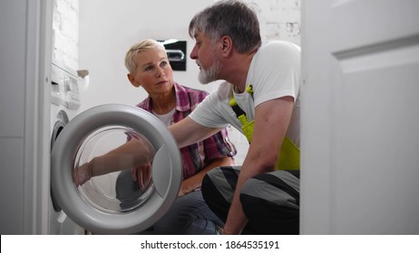 Aged Technician Examining And Repairing Tumble Dryer. Handyman Installing New Washing Machine And Showing Housewife How To Use It. Plumber Fixing Broken Washing Machine In Bathroom