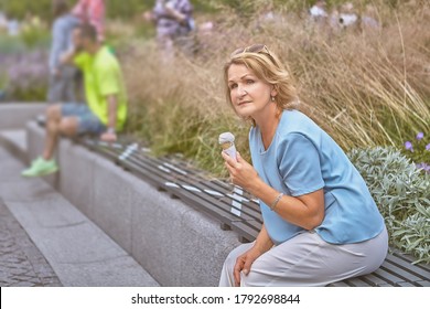 Aged White Attractive Lady  62 Years Old Is Sitting On The Bench With Social Distancing Markup In Public Park During Epidemic Of Covid-19 And Eating Ice Cream. She Has Casual And Elegant Cloth.