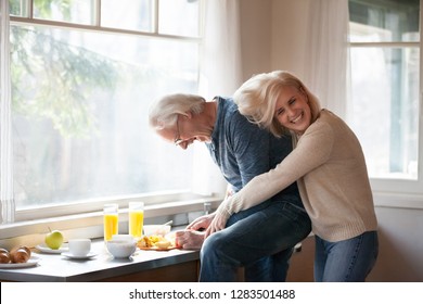 Aged wife husband in casual clothes preparing healthy breakfast standing in modern light kitchen, grey haired man cutting apples funny cheerful woman cuddle him from behind have fun laughing together - Powered by Shutterstock