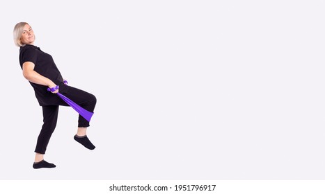 Aged Woman With Athletic Body Performs Exercises Using A Resistance Band. Photo Of Muscular Body Wearing Training Suit On Grey Background. Strength And Motivation. Full Length. High Quality Photo