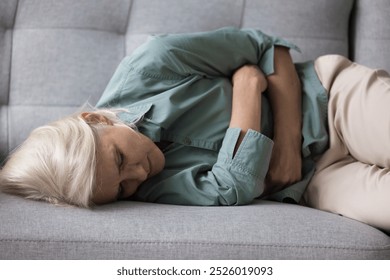 Aged woman clutching her stomach lying on sofa suffers from strong abdominal pain, chronic disease, discomfort, looks unhealthy, lack of energy or motivation, symptom of depression or physical ailment - Powered by Shutterstock