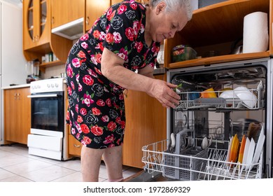 An Aged Woman In A Dressing Gown Unloads The Dishwasher In The Kitchen.