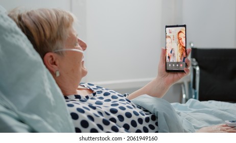 Aged woman holding smartphone with video call for remote communication with niece and daughter. Sick patient using online conference to talk to relatives while laying in hospital ward bed. - Powered by Shutterstock