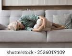 Aged woman lying on sofa, curled up, looks upset, dealing with mental pain or chronic illness, suffers from emotional distress, crisis or problems in personal life, experiencing anxiety and depression