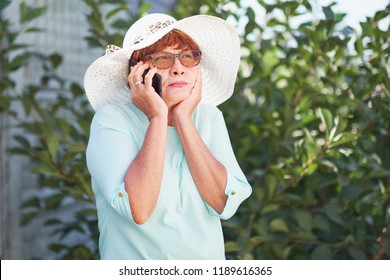 Aged Woman Talking On Phone. Old Lady In Park. Sunny Day, Happy Mood. Upset Senior Woman With Phone