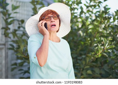 Aged Woman Talking On The Phone. Old Lady In Park. Sunny Day, Happy Mood. Upset Senior Woman With Phone