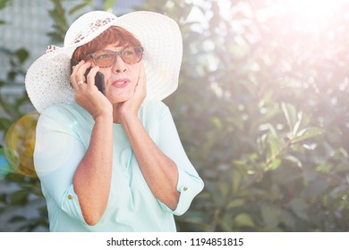 Aged Woman Talking On The Phone. Old Lady In Park. Sunny Day, Happy Mood. Upset Senior Woman With Phone