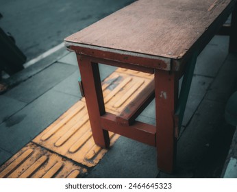 An aged wooden bench with a flat seat and no backrest. Its color appears to be faded red or brown. There is tactile paving, which is a textured ground surface used for visually impaired pedestrians. - Powered by Shutterstock