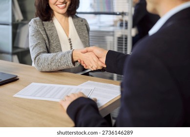 Agent and client handshake over contract in office. Smiling woman and man seal agreement and deal during a meeting. Forms and pen highlight contract signatures. Clear concept of business trust.