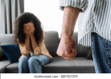Aggression by a man towards his woman, a man clenched his fist to hit his wife or girlfriend, the girl in defocus sitting on the couch covering herself with her hands, concept of domestic violence to