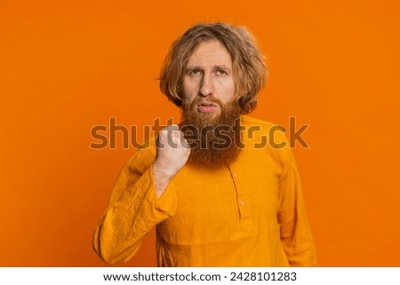 Aggressive angry Caucasian young man trying to fight at camera, shaking fist, boxing with expression, punishment, threaten, bullying, abuse, mad fury. Redhead guy isolated on orange studio background