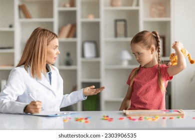 Aggressive Child Therapy. Cute Little Girl Feeling Angry During Therapy Session With Psychotherapist Lady, Upset Small Kid Threaten Specialist Doctor With A Toy, Having Temper Tantrum