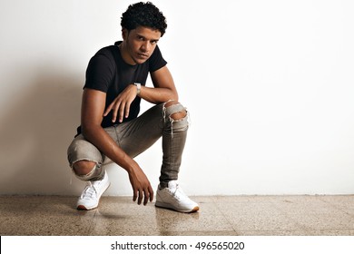 Aggressive and gloomy looking attractive black model in slim torn grey jeans and plain black t-shirt crouching at a white wall