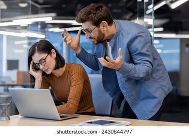 Aggressive male boss in denim shirt spreading hands and yelling at annoyed female employee by working desk. Stressed woman sitting at office in front of laptop and taking critique from director. - Powered by Shutterstock