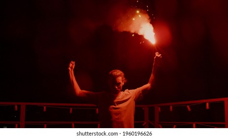 Aggressive man burning signal flare on night roof. Serious football fan standing with red fire outdoors. Man protest concept. Male protester with light at urban background.