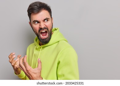 Aggressive outraged bearded European man gestures angrily screams loudly expresses rage hates something turns back wears casual hoodie poses against grey background blank space for your text - Shutterstock ID 2033425331