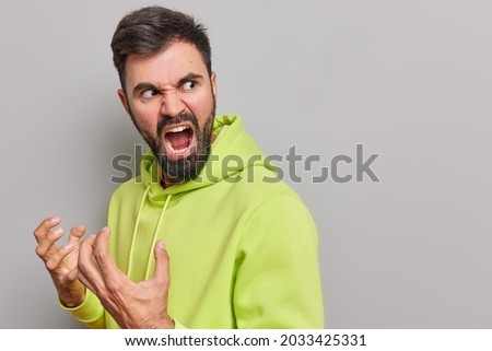 Aggressive outraged bearded European man gestures angrily screams loudly expresses rage hates something turns back wears casual hoodie poses against grey background blank space for your text