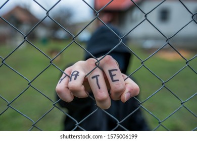 Aggressive Teenage Boy Holding The Wired Fence At The Correctional Institute, The Word Hate Is Written On Hes Hand, Conceptual Image Of Juvenile Delinquency .