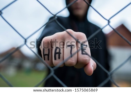 Aggressive teenage boy showing hes fist behind wired fence at the correctional institute, the word hate is written on hes hand, focus on the boys hand , conceptual image of juvenile delinquency .