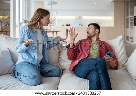 Aggressive wife with husband trying to talk gesturing hands. Family couple having misunderstanding showdown scandal sitting on couch at home. Neurotic relationships, toxic relations, marital discord.