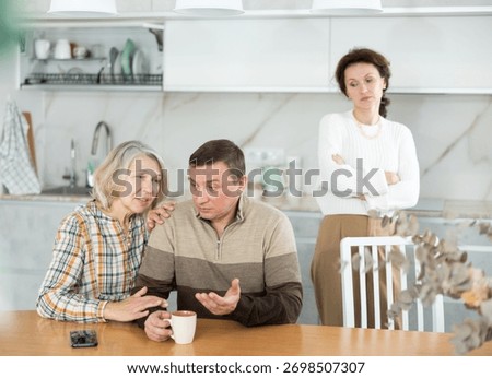 Aggrieved middle-aged man sitting at the kitchen-table after brawling with wife and old woman speaking to him peacefully
