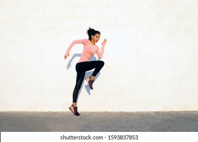 Agile and fit young woman jumping and running on the white concrete wall background, facing right, wearing black sport tights and pink shirt - Shutterstock ID 1593837853