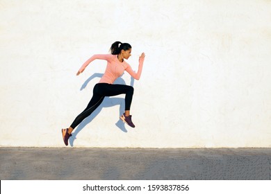 Agile And Fit Young Woman Jumping And Running On The White Concrete Wall Background, Facing Right, Wearing Black Sport Tights And Pink Shirt