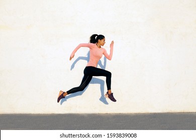 Agile And Fit Young Woman Jumping And Running On The White Concrete Wall Background, Facing Right, Wearing Black Sport Tights And Pink Shirt