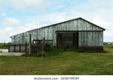 An aging barn with a rustic charm sits amidst lush green fields, embodying rural life and agricultural heritage, under a bright cloudy sky on a peaceful day.