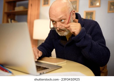 Aging and eyesight problems. Side view image of senior bald bearded grandfather in elegant stylish blue cardigan using laptop in his cabinet, reading article, adjusting glasses on his nose