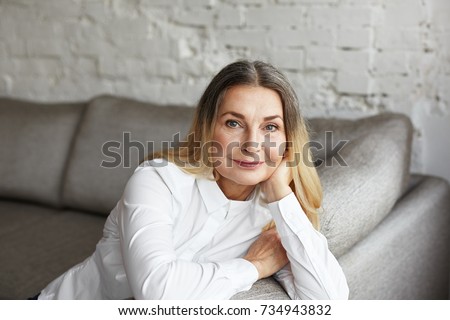 Aging gracefully. Indoor portrait of attractive mature 60 year old female with beautiful blue eyes and healthy wrinkled skin smiling happily at camera while relaxing on grey comfortable sofa at home