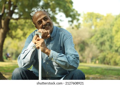 Aging Man Having Joint Pain In His Knee In A Lush Green Park In Noon. Elderly Retired Man Holding A Walking Stick. Showcasing Health Care Osteoarthritis Concept. Old Man Showing Various Expressions.