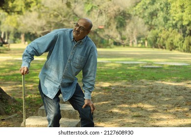 Aging Man Having Joint Pain In His Knee In A Lush Green Park In Noon. Elderly Retired Man Holding A Walking Stick. Showcasing Health Care Osteoarthritis Concept. Old Man Showing Various Expressions.