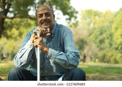 Aging Man Having Smiles In A Lush Green Park In Afternoon. Elderly Retired Man Holding A Walking Stick. Showcasing Old Age Concept.