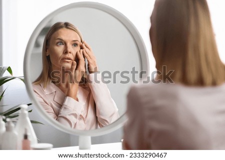 Aging Signs. Upset mature woman looking at wrinkles around her eyes while standing near mirror at home, beautiful middle aged lady inspecting face, feeling stressed, selective focus