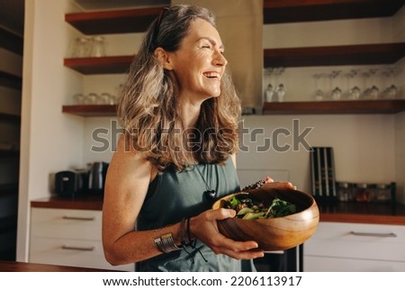Aging woman smiling happily while holding a buddha bowl in her kitchen. Happy senior woman serving herself a healthy vegan meal at home. Mature woman taking care of her body with a plant-based diet.