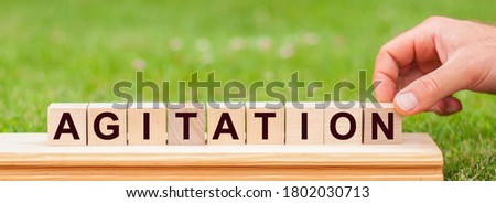AGITATION word made with building blocks. Man hand holding wooden cube block with AGITATION business word on green lawn background.
