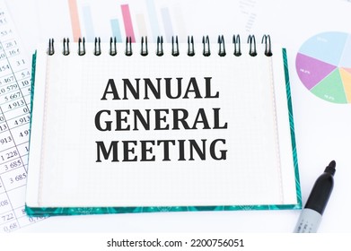 AGM Annual General Meeting is written in red on a white piece of paper on a light yellow background next to a laptop, pen, magnifying glass, glasses and a green plant. - Shutterstock ID 2200756051