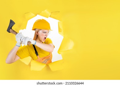 Agressive repair woman in helmet, uniform is holding metal ax in torn hole of yellow background. Instruments, tools, accessories for renovation, building house. Female worker is working at hard job. 
