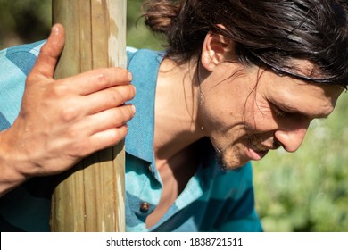 An Agricultor Wearing A Stripped Blue Shirt, Working On His Orchard. He Is Looking Down At The Crops While Holding A Wooden Post With His Right Hand. He Has Messy Long Hair And A Pony Tail 