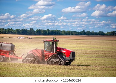 Agricultural Background With Red Tractor Pulling Plow, Throwing Dust In Air. Combine Harvester At Wheat Field. Heavy Machinery During Cultivation, Working On Fields. Dramatic Sky, Rain, Storm Clouds
