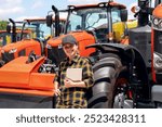 Agricultural or Construction equipment dealer. Woman in plaid shirt and cap with digital tablet in her hand leaned on the wheel of the new tractor.