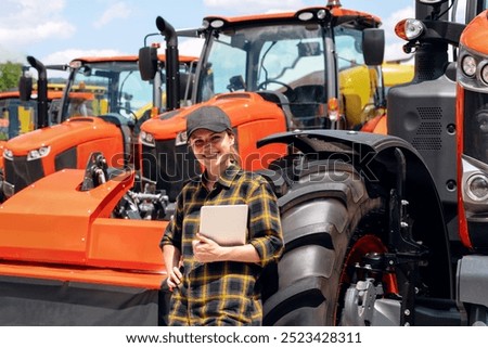 Agricultural or Construction equipment dealer. Woman in plaid shirt and cap with digital tablet in her hand leaned on the wheel of the new tractor.