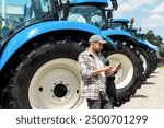 Agricultural equipment dealership. Agribusiness. Man leaned on a tractor wheel and using digital tablet.