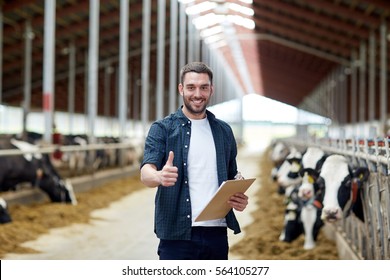 Agriculture Industry, Farming, People And Animal Husbandry Concept - Happy Smiling Young Man Or Farmer With Clipboard And Cows In Cowshed On Dairy Farm Showing Thumbs Up Hand Sign