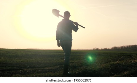 An Agronomist Man Walks Through A Black Plowed Field At Sunset, Farmer With A Shovel In His Hands, Work In The Field For The Worker, Dawn Time In The Sky For Life Activities In Agriculture