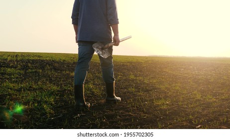 An agronomist man walks through a black plowed field at sunset. Farmer with a shovel in his hands.