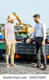 Agronomist With Salesman Choosing A New Farm Truck Trailer, Standing On The Open Ground Of The Agricultural Shop